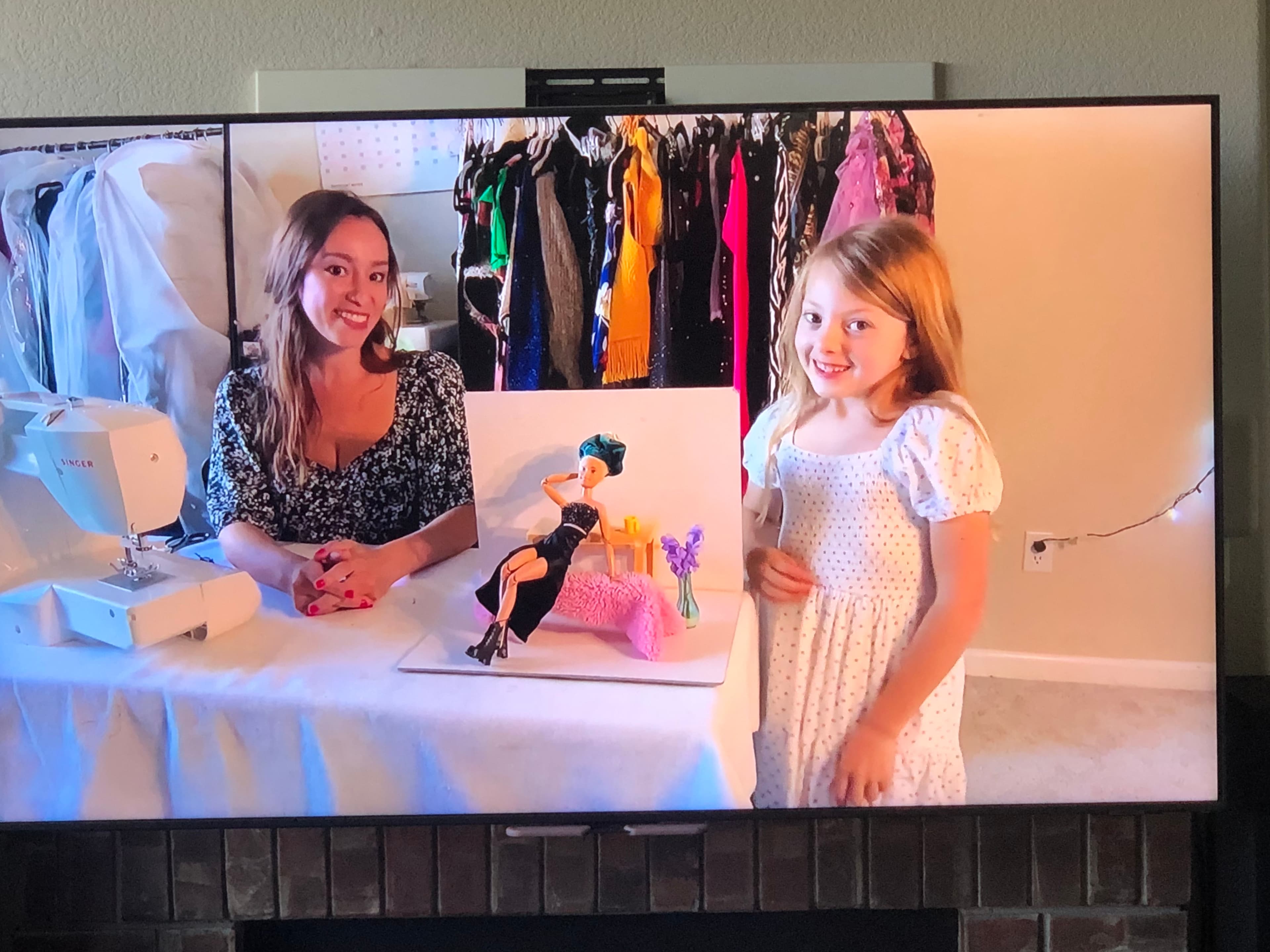 A sewing scene with two people at a table with a Singer machine, a fashion doll display, and clothing racks visible in background. One wears floral, other in white dress.