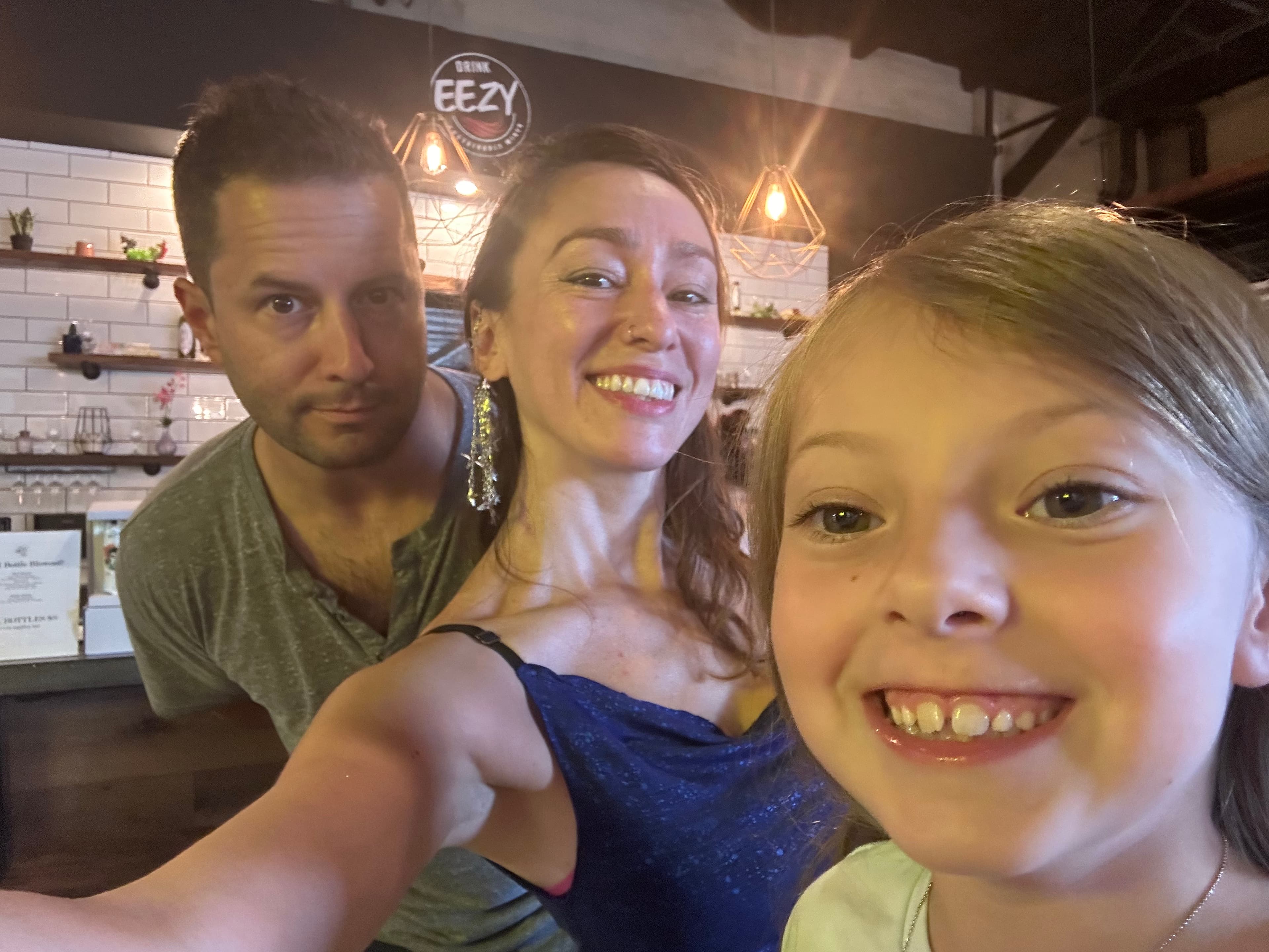 A family selfie taken at a restaurant or bar called "Eezy", with three people smiling at the camera. The interior has white brick walls and hanging geometric lights.