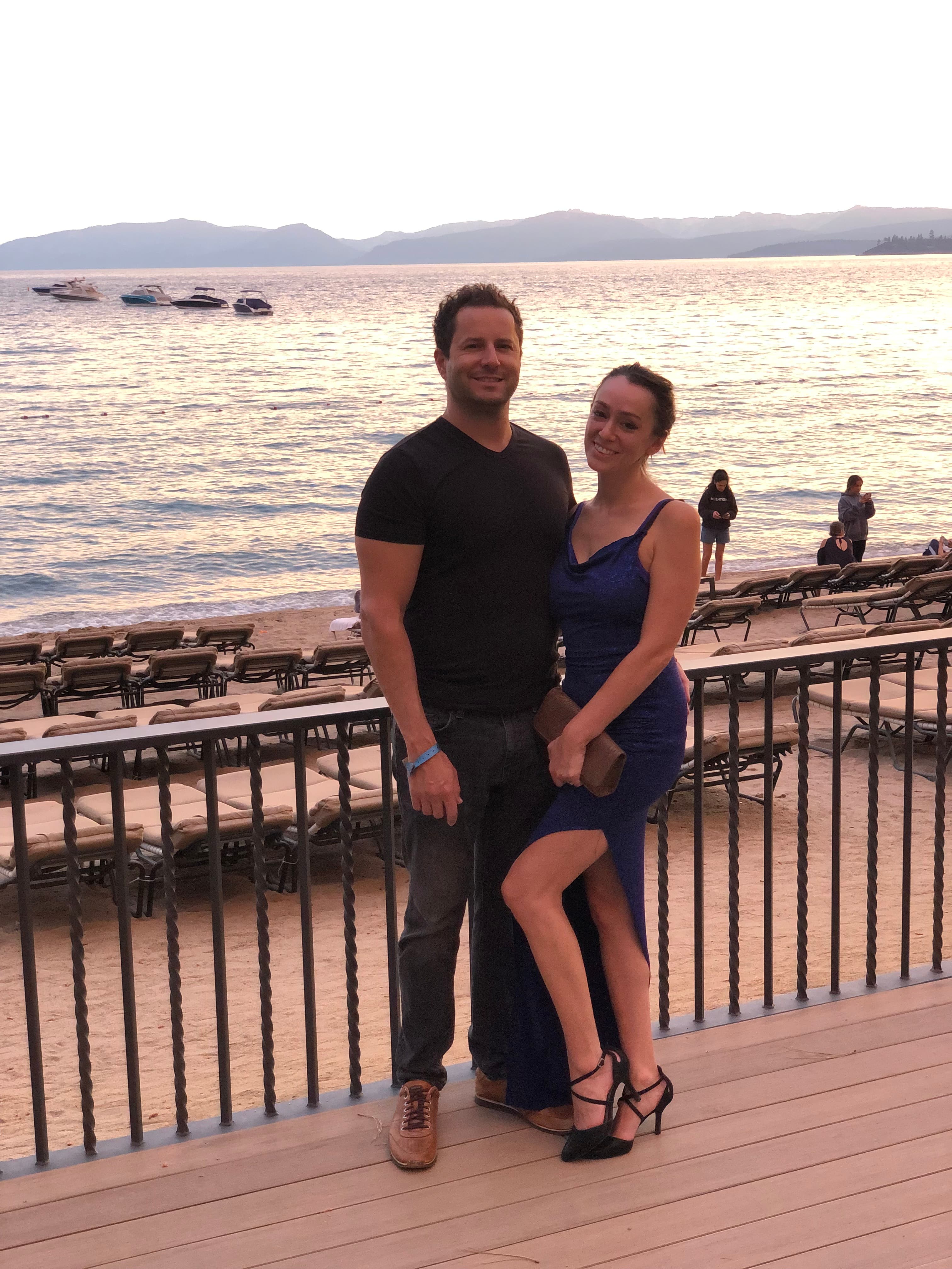 A couple poses on a lakeside deck at sunset, with boats in the water and mountains in background. She wears a blue dress, he wears black, with beach chairs behind them.
