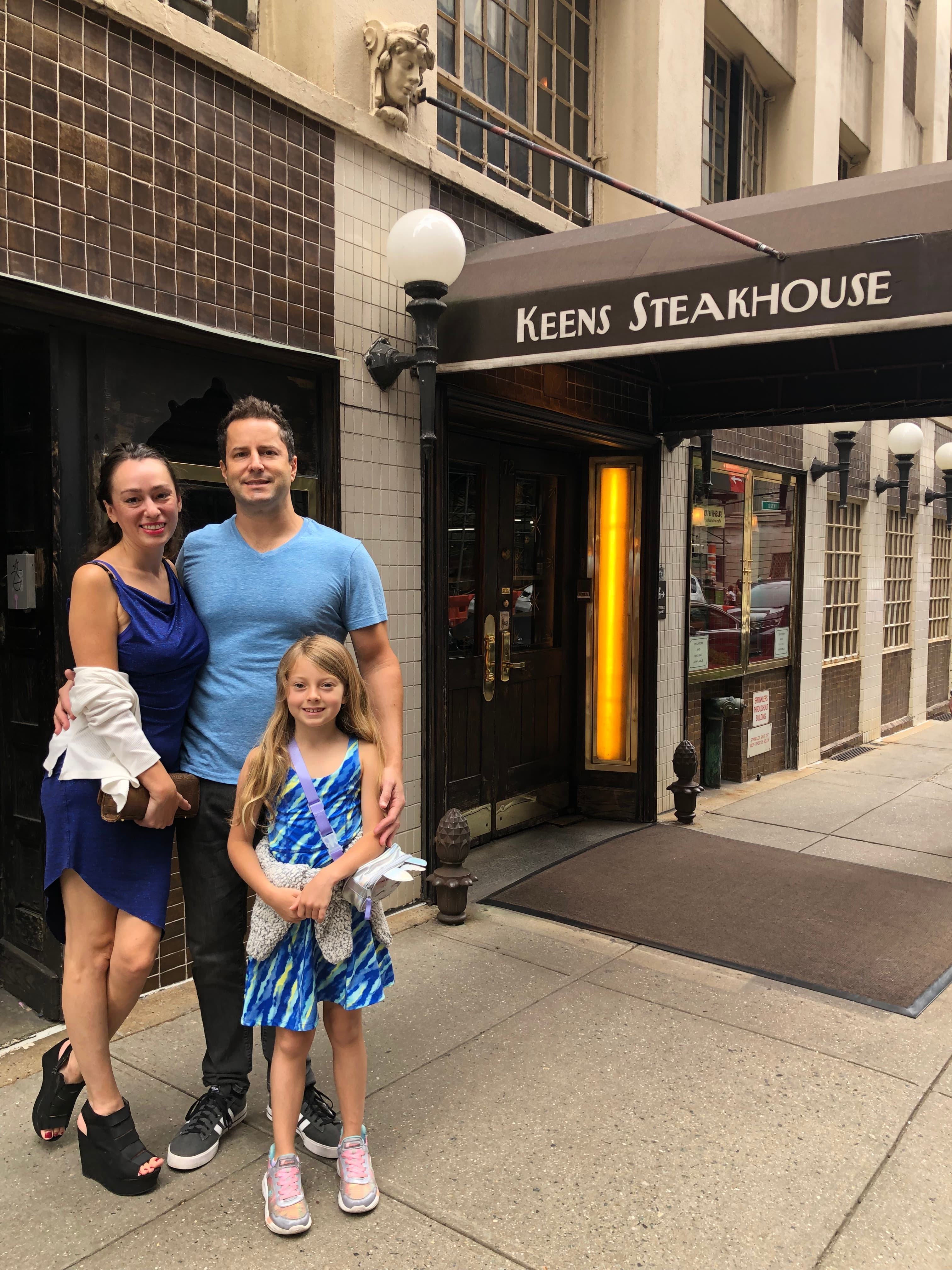 A family of three stands outside Keens Steakhouse in New York City, wearing coordinating blue outfits on a cloudy day.