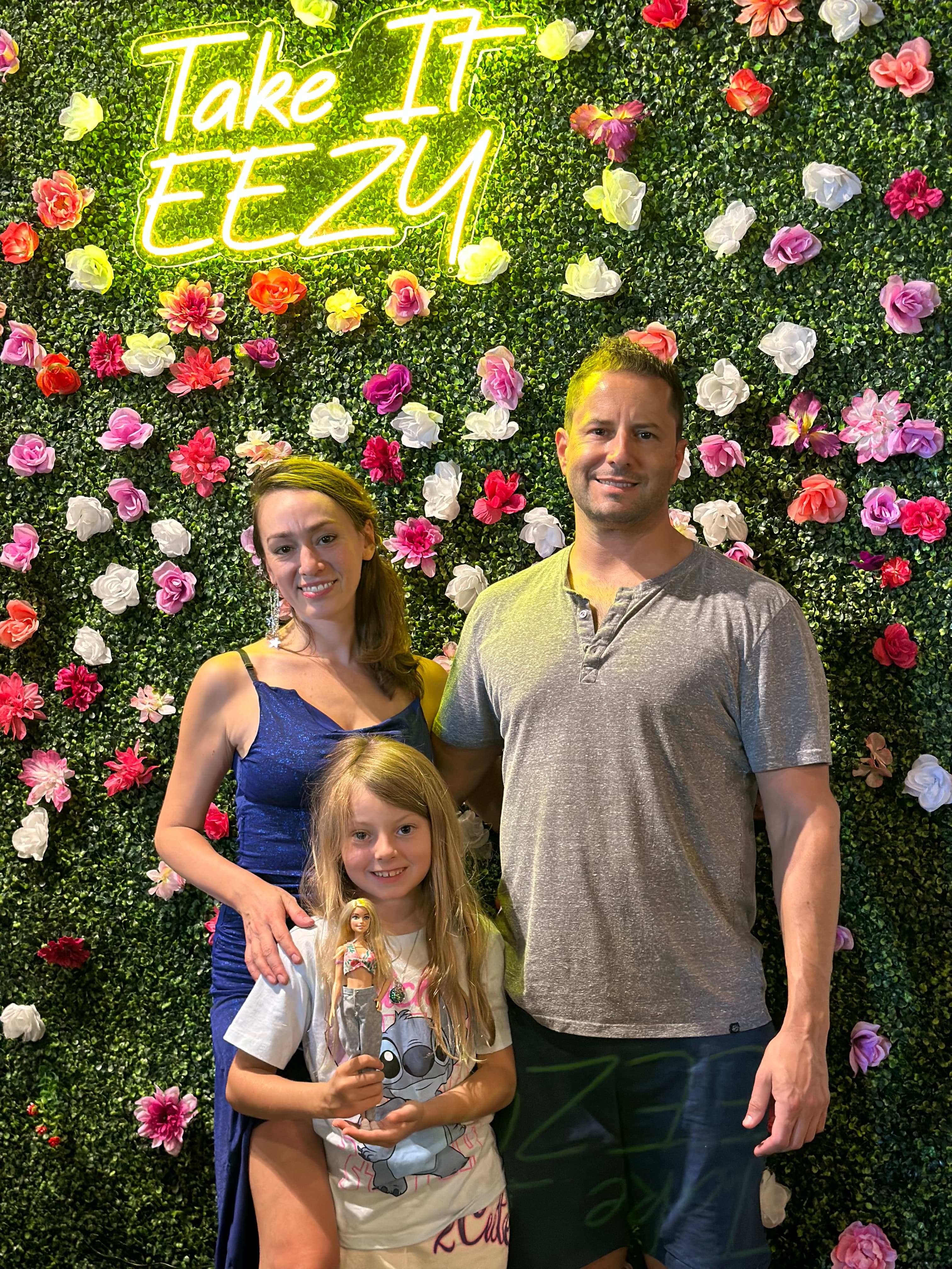 Photo of a family in front of a floral wall display with yellow neon "Take It Eezy" sign. Young girl holds Barbie doll. Mom wears blue dress, dad in grey shirt.