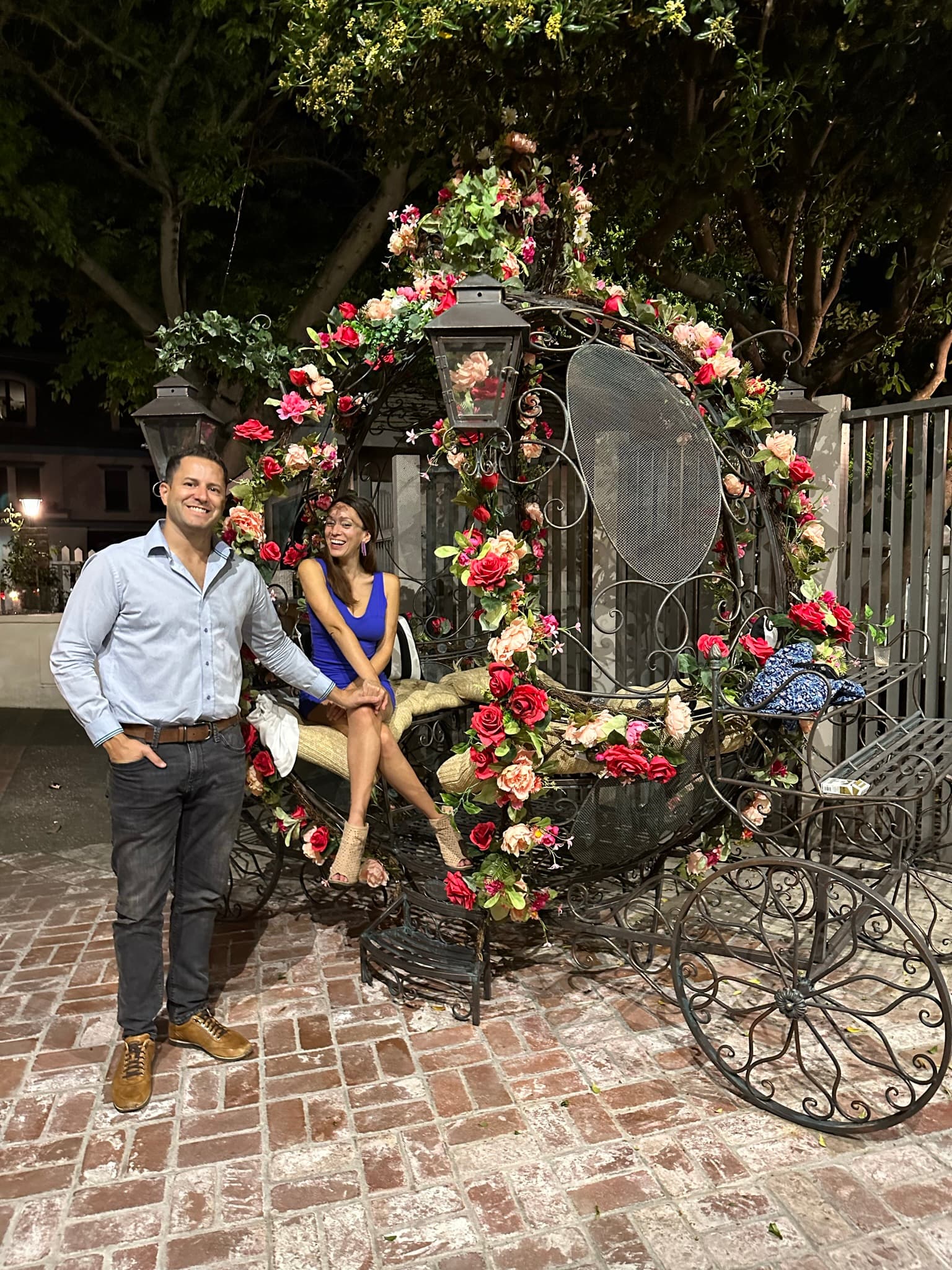 A couple poses with a decorative metal carriage covered in red and pink roses, with lanterns, on a brick patio at night.