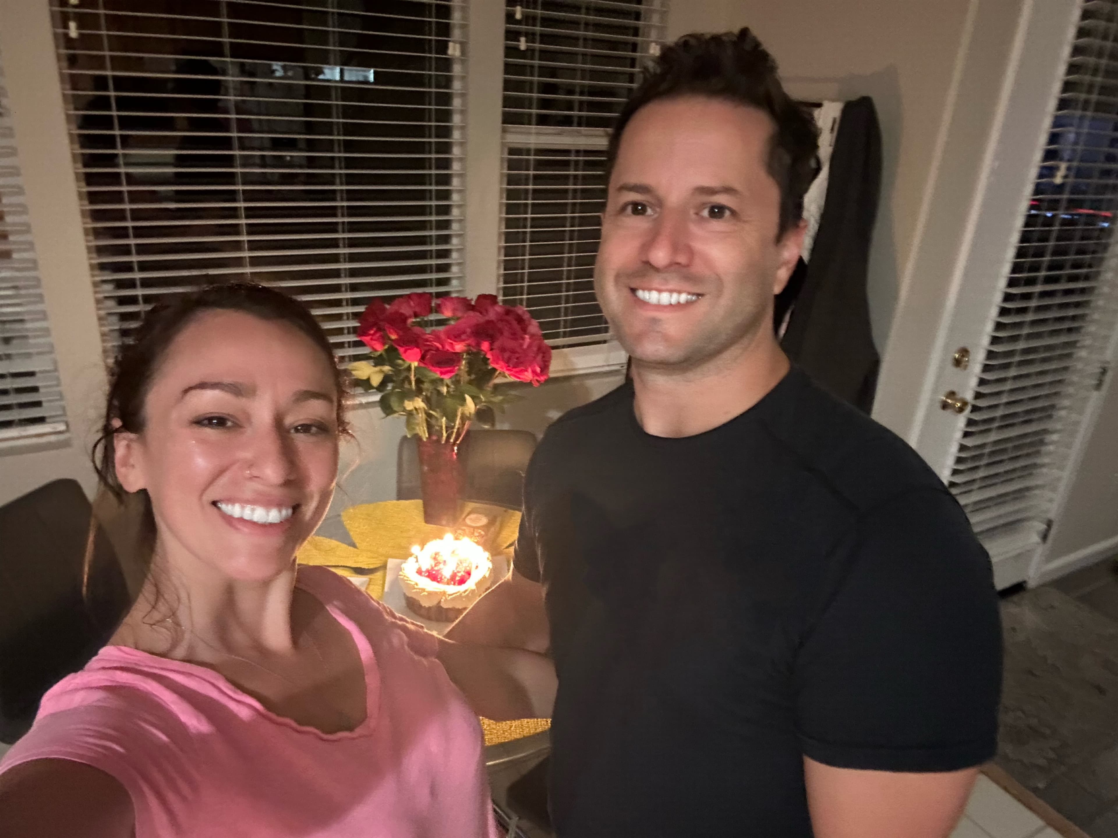 A couple smiling at camera in a dimly lit room with a lit birthday cake and red roses in a vase on a table behind them. Woman in pink, man in black shirt.