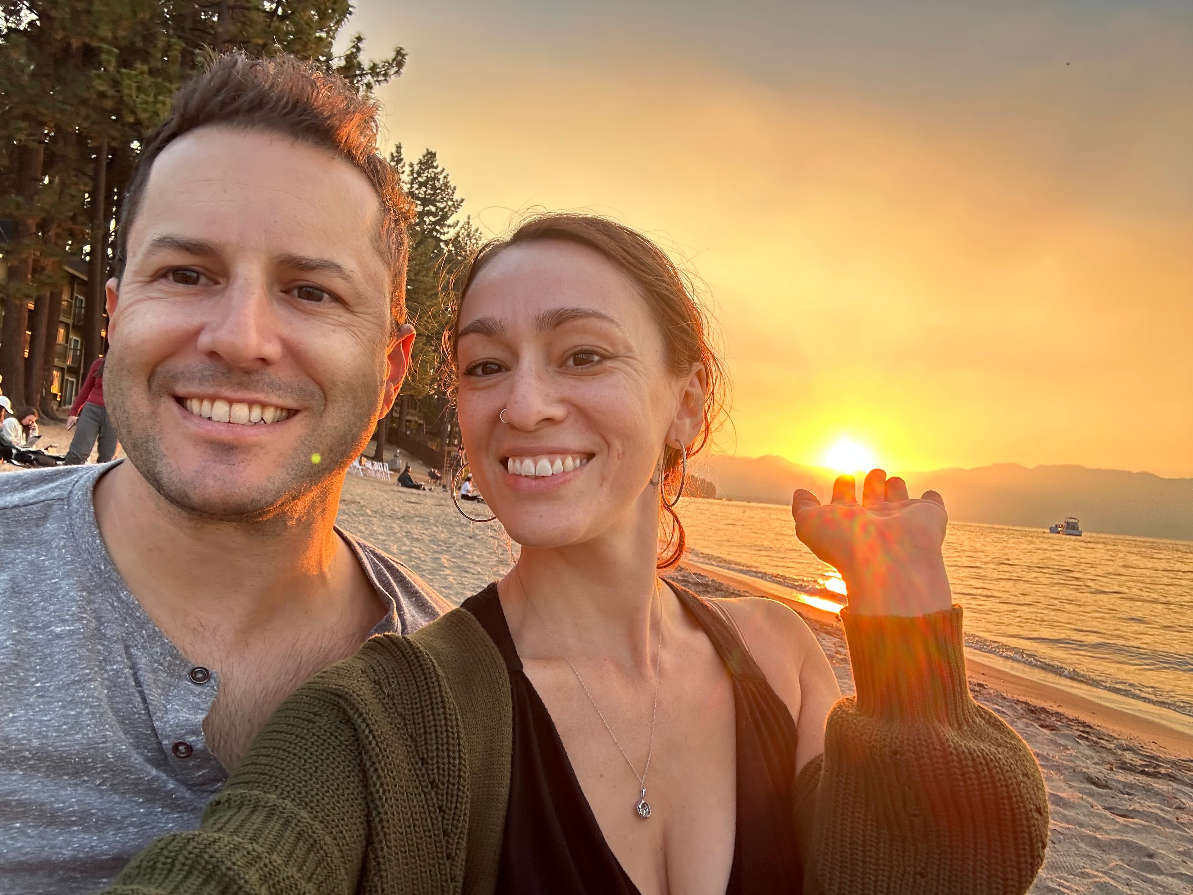A couple takes a selfie on a beach at sunset, with golden light and mountains in the background. Pine trees line the shore.