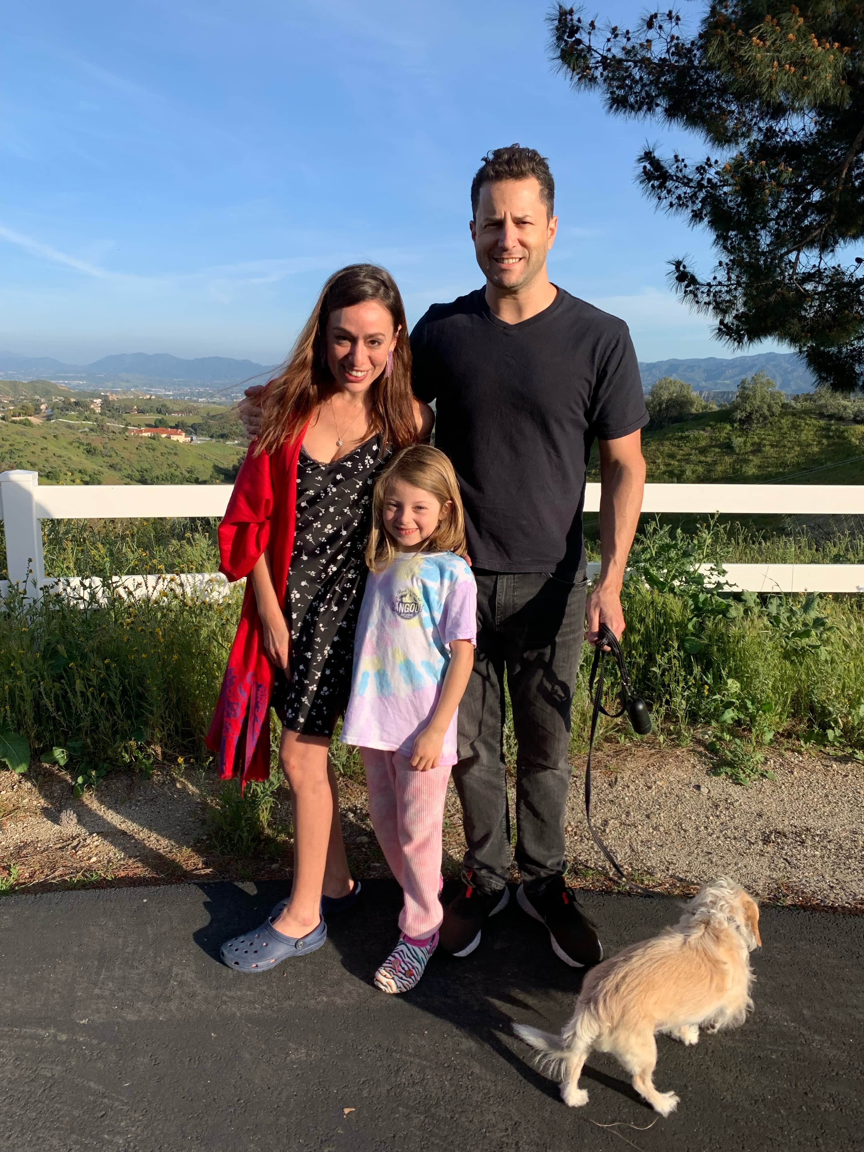 A family of three standing outdoors with their dog against a white fence and mountain vista backdrop. Mother wears red cardigan, child in tie-dye, father in black shirt.