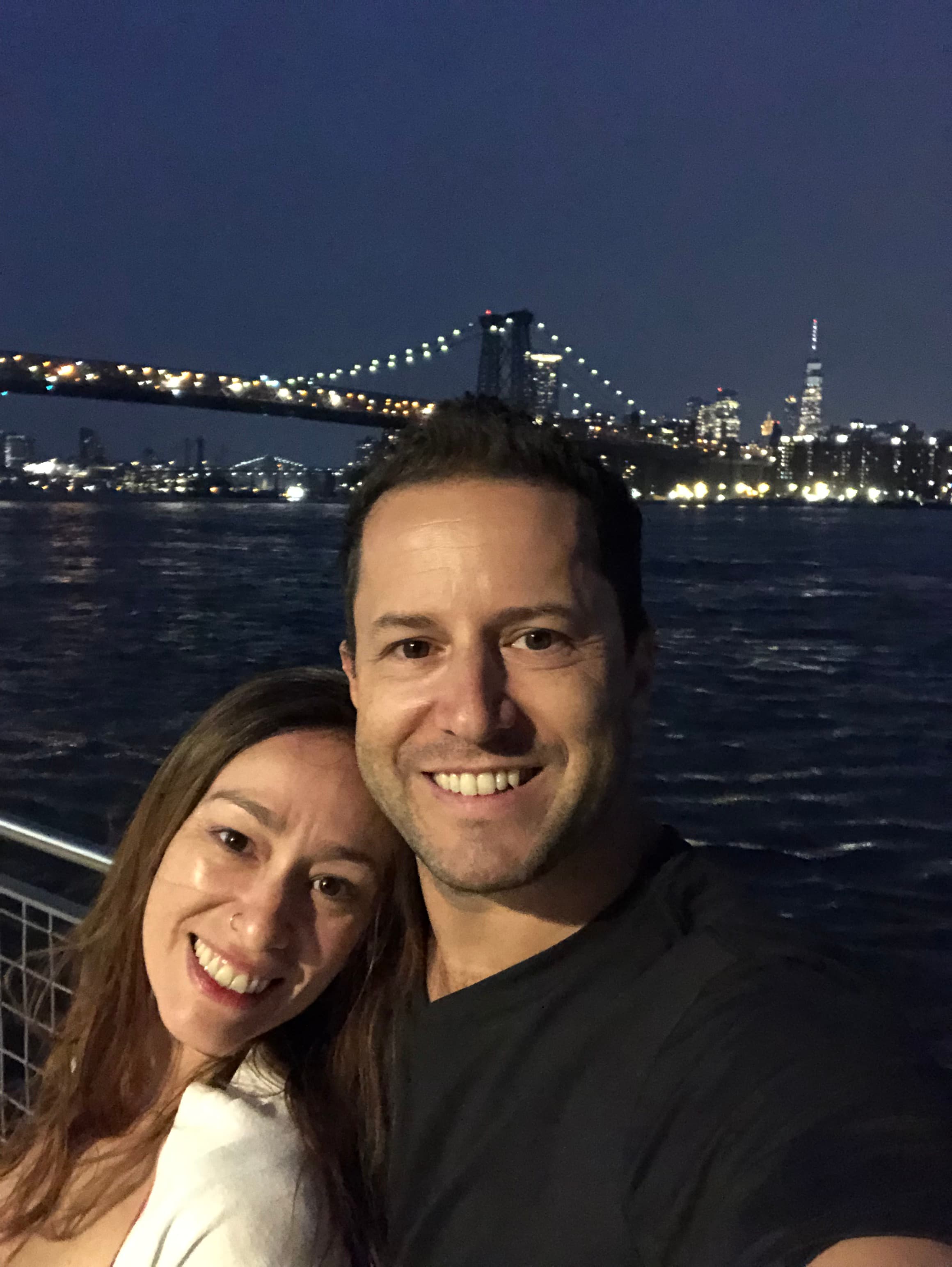 A couple takes a selfie at night with the illuminated Manhattan Bridge and NYC skyline visible in the background over dark waters.