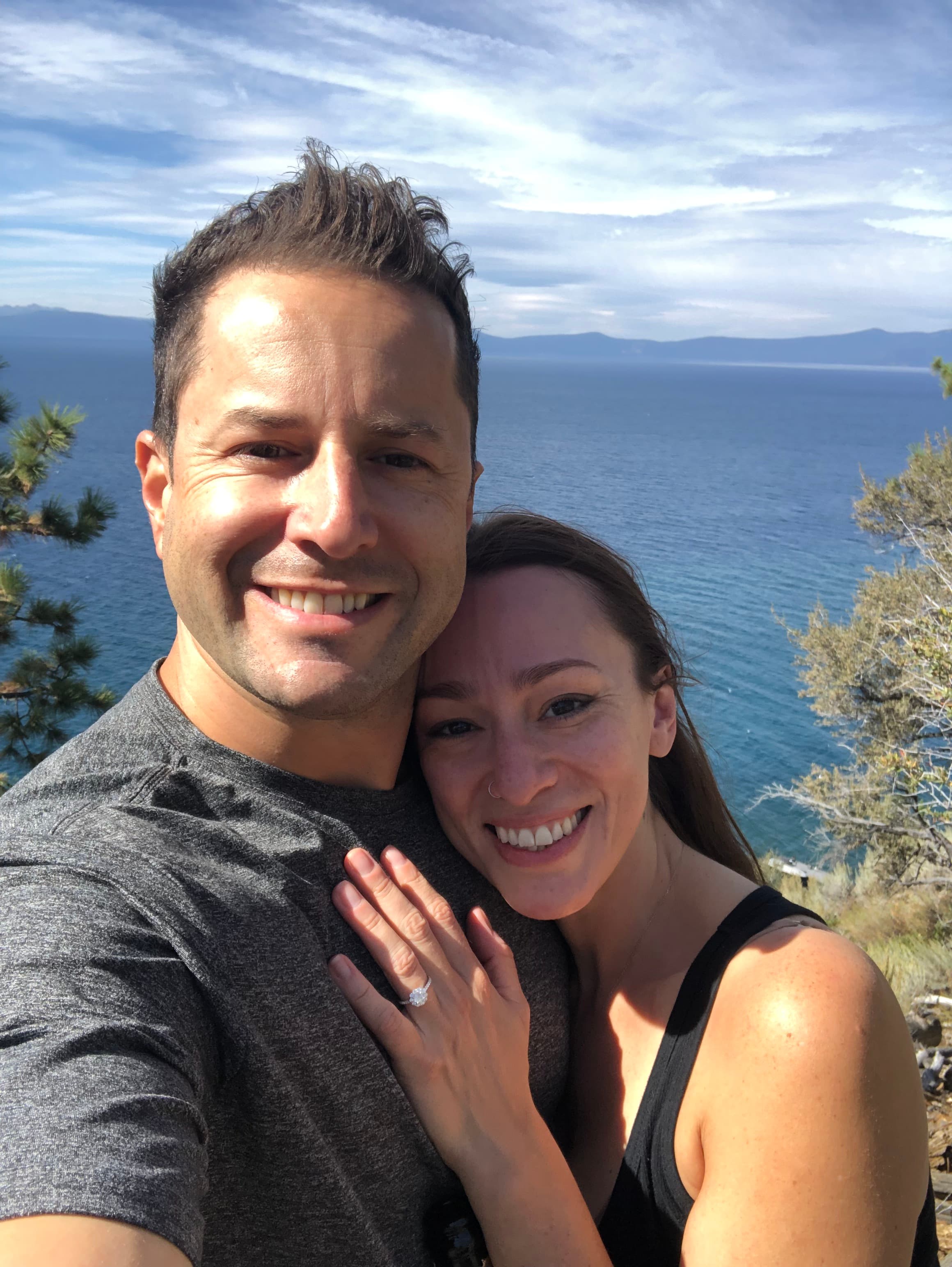A couple takes a selfie overlooking Lake Tahoe, with mountains and blue water in the background. The woman displays an engagement ring.