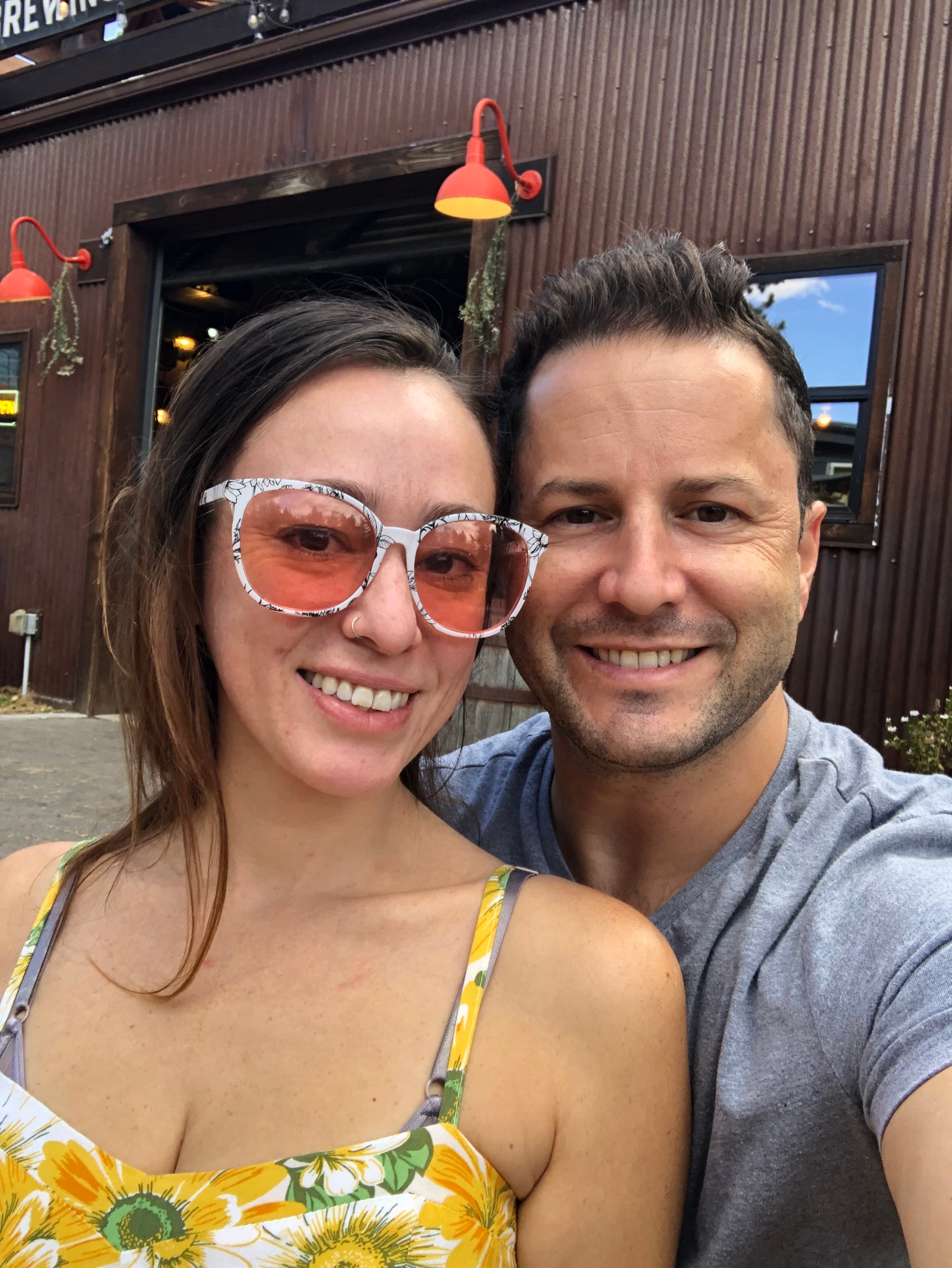 A smiling couple takes a selfie outside a restaurant with brown walls and orange lamps. She wears sunglasses and a floral top, he wears a grey shirt.