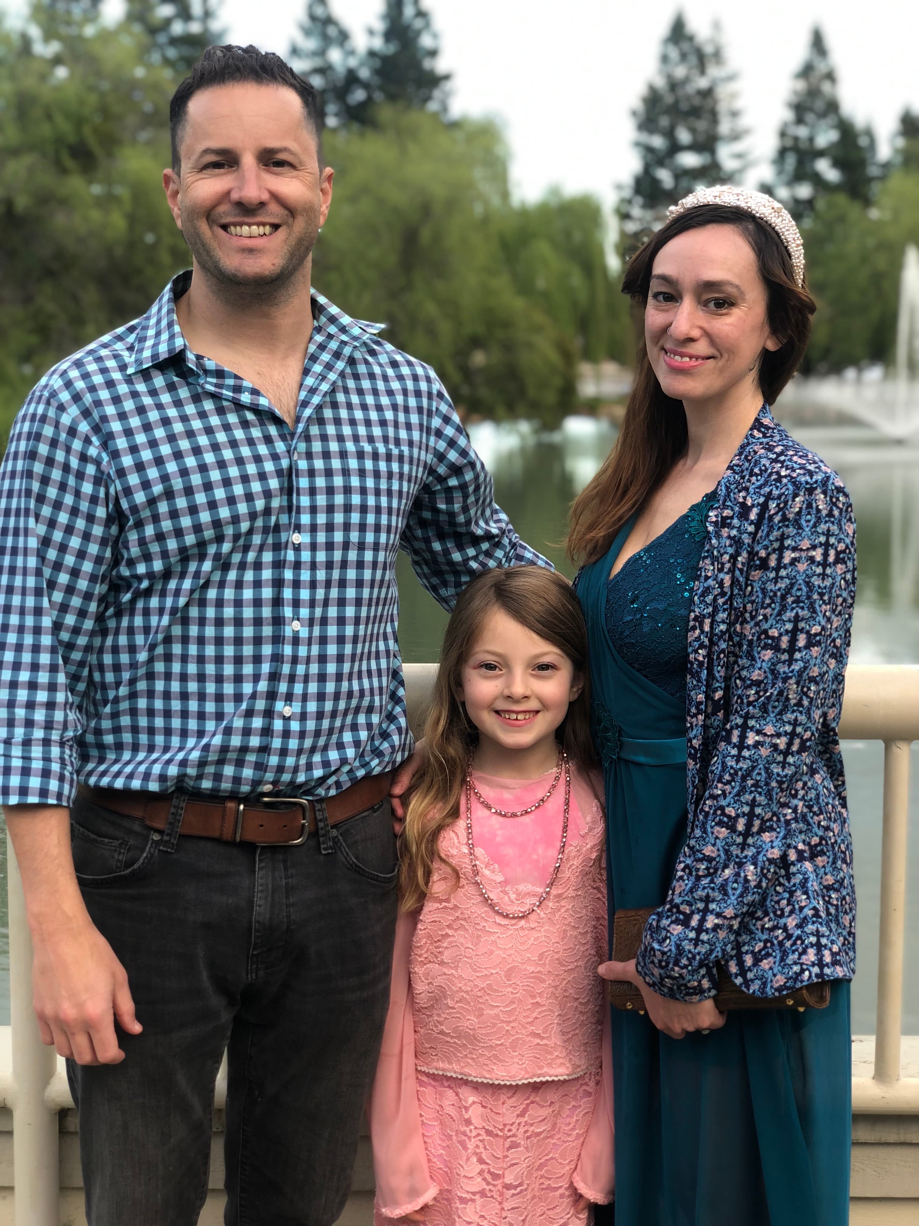 A family of three poses outdoors by a lake. The father wears a checkered shirt, the mother wears a blue dress, and their young daughter wears a pink lace dress.