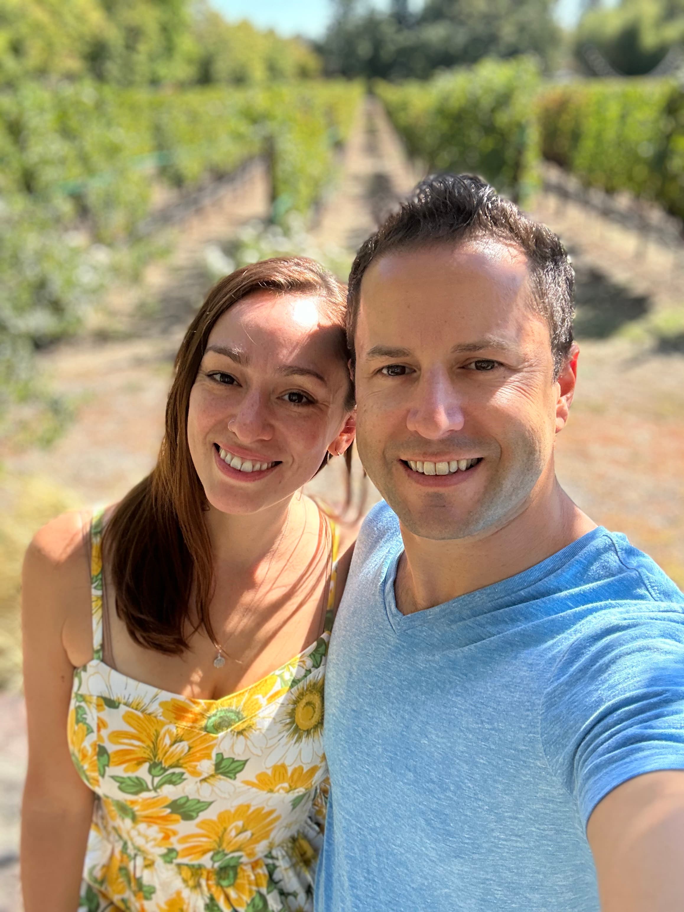 A couple takes a selfie in a vineyard, wearing summer attire - she in a sunflower print dress and he in a blue t-shirt. Grapevines visible in background.